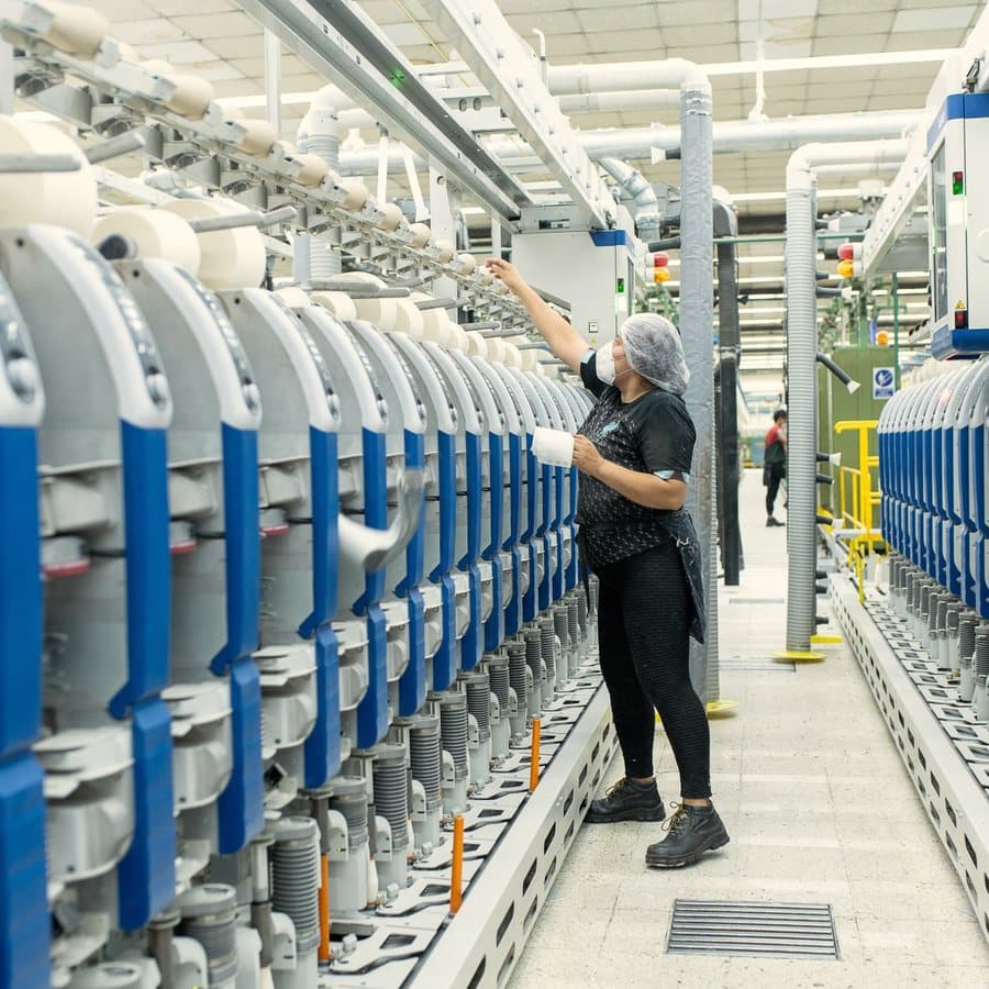 Sala de hilado de la planta con filas de máquinas continuas y conos de hilo crudo.